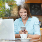 mujer sonriendo revisando el móvil en una cafetería frente a un portatil